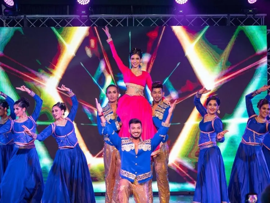 Divaksha performing with two dancers under stage lighting, marigold columns behind