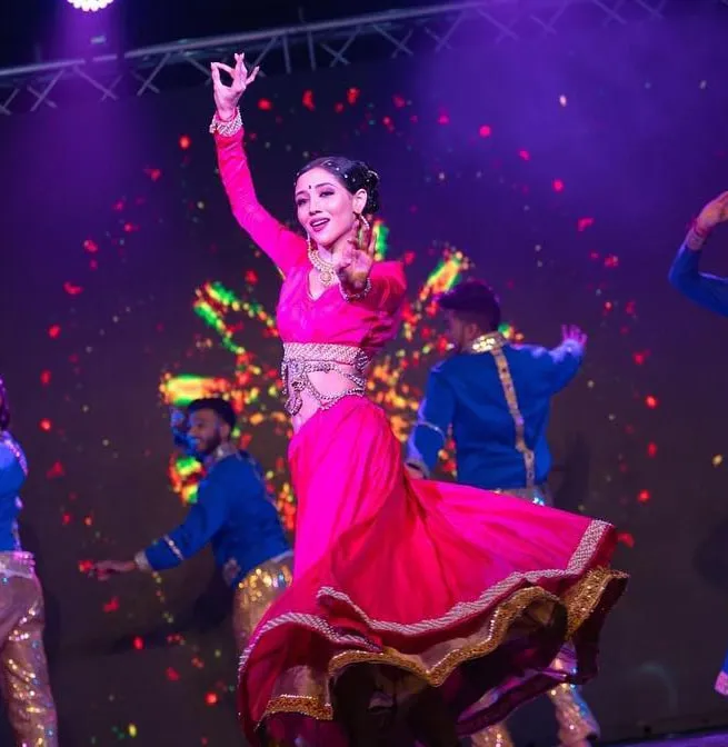 Divaksha mid-turn in a pink lehenga under stage lighting, one arm raised