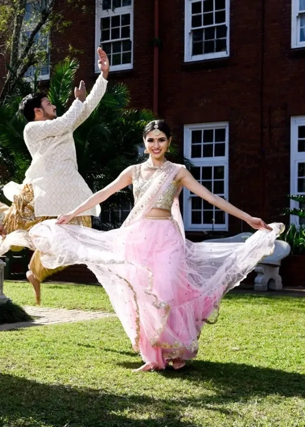 Divaksha Jinabhai turning in a pale pink lehenga, holding the skirt wide, on a lawn in front of a red brick building