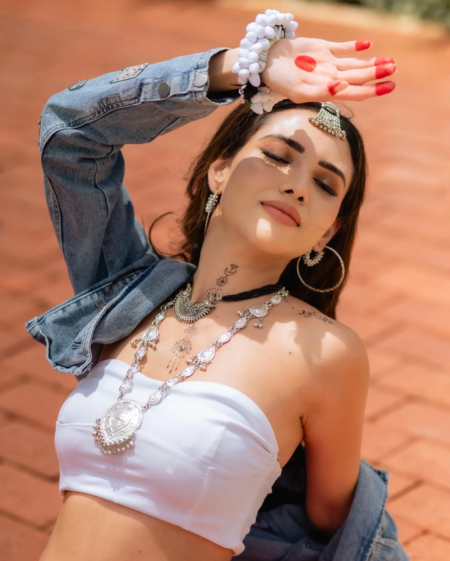 Divaksha Jinabhai in sunlight with her eyes closed, one hand shading her face, in a white top and silver necklaces