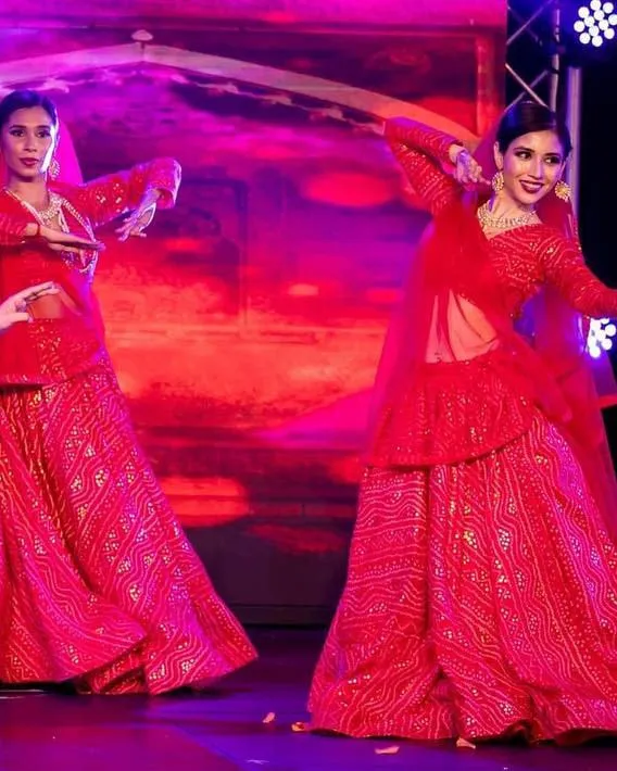Divaksha in red mid-phrase on a lit stage, another dancer behind her