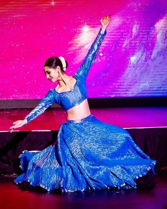 Divaksha in a blue sequinned lehenga, kneeling mid-phrase with one arm raised, against a lit stage screen