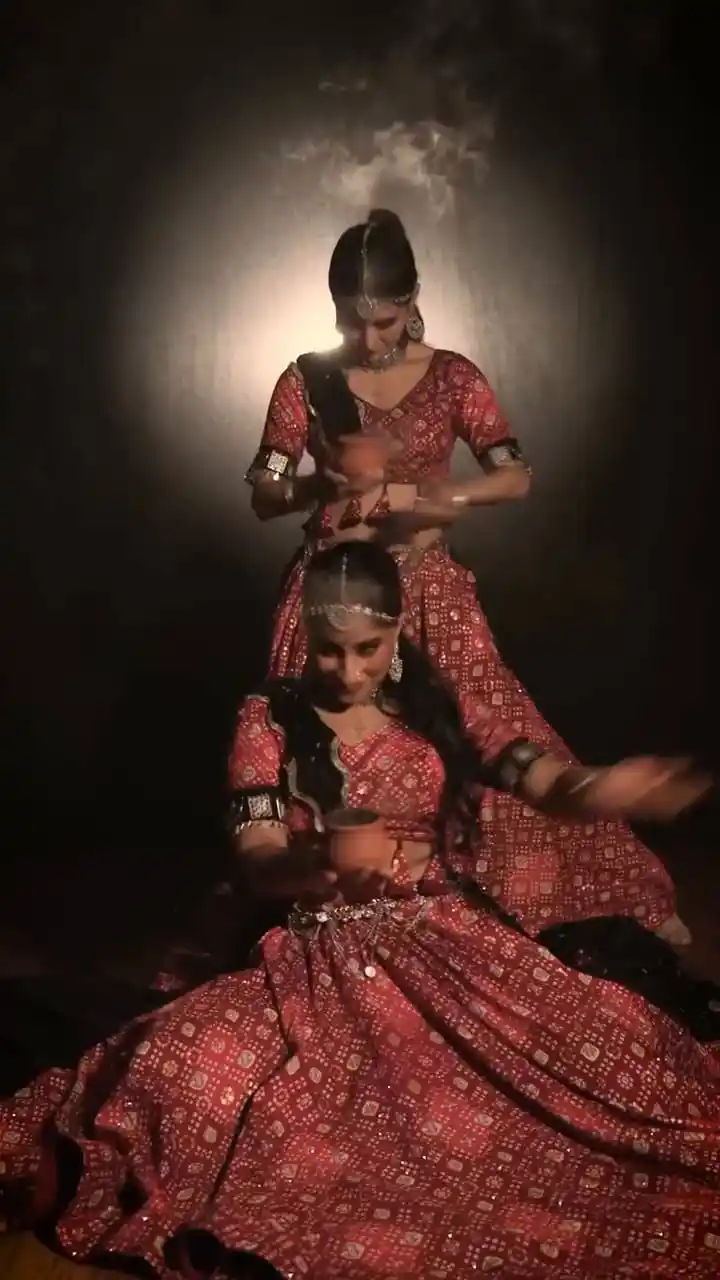 Divaksha Jinabhai and Primeshni Govender in red bandhani lehengas, holding clay pots under a single spotlight