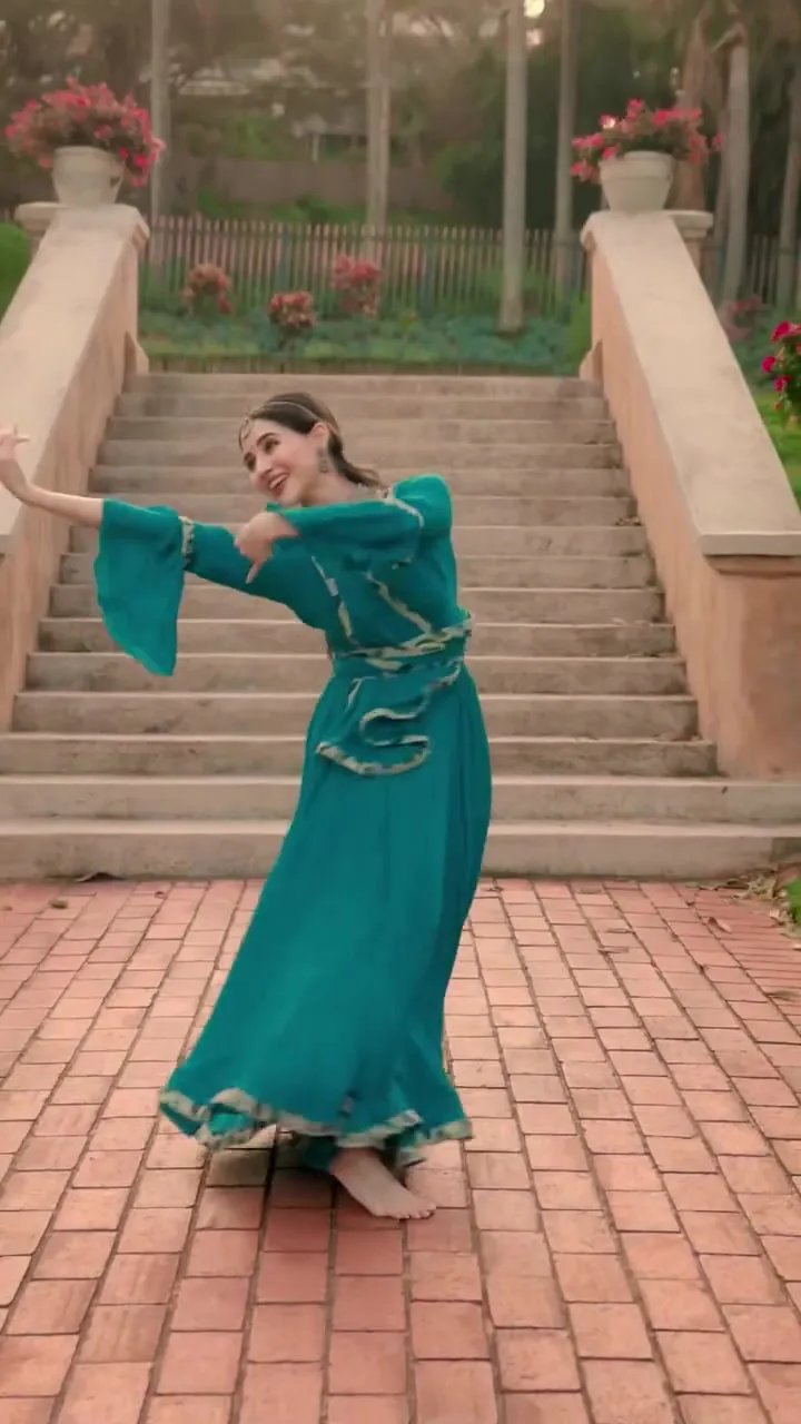 Divaksha in a teal anarkali on a brick path, arms extended mid-phrase